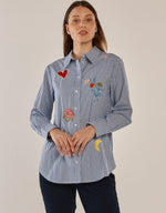 Load image into Gallery viewer, A woman modeling a light blue and white vertically striped button-up shirt with colorful embroidered patches, including a red heart, a flower, and a crescent moon, paired with dark pants against a plain beige background.

