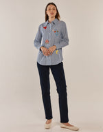 Load image into Gallery viewer, A woman modeling a light blue and white striped button-down shirt with colorful embroidered patches, paired with dark navy pants and beige flats, against a plain off-white studio background.

