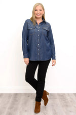 Load image into Gallery viewer, A woman modeling a blue denim long-sleeve western-style shirt with chest pockets, paired with black pants and brown ankle boots, standing against a white wall and light wood floor.
