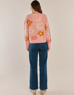 Load image into Gallery viewer, A woman viewed from behind wearing a pink knit sweater with large orange and cream flower motifs, paired with blue jeans and light-colored sneakers, against a plain light beige studio background.
