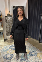 Load image into Gallery viewer, A woman stands in a boutique fitting room wearing a black long-sleeve knit top and matching black midi skirt, with gray pointed-toe boots. A mannequin in a patterned dress and a mirror are visible in the background, along with dark gray curtain doors and a blue and yellow patterned rug.
