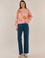 Load image into Gallery viewer, A woman modeling a pink crewneck sweater with a retro orange and yellow flower pattern, paired with blue jeans and beige sneakers, against a plain beige studio background.
