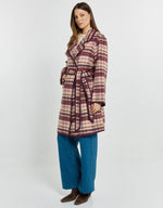 Load image into Gallery viewer, A woman modeling a long plaid wrap coat in burgundy, beige, and brown tones, paired with wide-leg blue jeans and brown ankle boots, against a plain light gray studio background.
