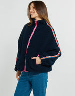 Load image into Gallery viewer, A woman wearing a navy blue sherpa fleece zip-up jacket with bright pink and orange striped accents on the zipper and sleeves, paired with blue jeans, against a light gray studio background.
