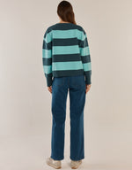 Load image into Gallery viewer, A woman viewed from behind wearing a long-sleeve knit sweater with horizontal dark teal and mint green stripes, paired with blue jeans and white sneakers, against a plain light beige studio background.
