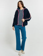 Load image into Gallery viewer, A woman modeling a navy blue sherpa zip-up hoodie jacket with pink and white striped trim, worn over a navy and white striped t-shirt, blue jeans, and white sneakers, against a light gray studio background.
