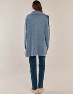 Load image into Gallery viewer, A woman viewed from behind wearing a cornflower blue knit cardigan over dark blue jeans and cream-colored slip-on shoes, set against a plain light beige studio background.
