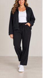 Load image into Gallery viewer, A woman modeling a black zip-up jacket and matching black drawstring sweatpants over a white tank top and white sneakers, photographed against a plain light beige studio background.
