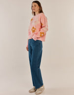 Load image into Gallery viewer, A woman modeling a pink crewneck sweater with yellow and orange floral patterns, paired with blue jeans and beige sneakers, against a plain light beige studio background.
