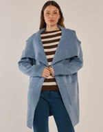 Load image into Gallery viewer, A woman modeling a cornflower blue open-front knit cardigan over a brown and cream striped sweater, paired with dark blue jeans, against a plain light beige studio background.

