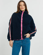 Load image into Gallery viewer, A woman wearing a navy blue sherpa fleece zip-up jacket with bright pink and white striped details on the sleeves, paired with blue jeans, against a light gray studio background.
