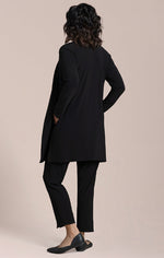 Load image into Gallery viewer, A woman shown from the back wearing a black long-sleeve tunic-style cardigan, black cropped pants, and black flats against a plain warm beige studio background.
