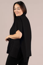 Load image into Gallery viewer, A woman modeling a black short-sleeve cape-style top with a high mock neck, photographed against a solid light beige studio background.
