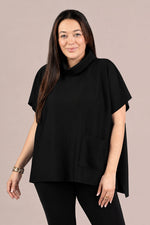 Load image into Gallery viewer, A woman modeling a black short-sleeve turtleneck poncho top with a front patch pocket, paired with black leggings, against a solid light taupe background.
