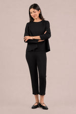 Load image into Gallery viewer, A woman modeling a black two-piece outfit consisting of a long-sleeve peplum-style top and matching cropped pants, paired with black flats, against a solid light beige studio background.
