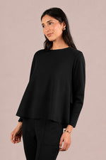 Load image into Gallery viewer, A woman modeling a black long-sleeve top with a boat neckline and a draped, asymmetrical hem, paired with black trousers, against a solid light beige background.
