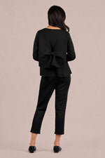 Load image into Gallery viewer, A woman shown from the back wearing a black long-sleeve top with a large bow detail at the back neckline and black cropped pants, standing against a plain warm beige studio background.
