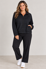 Load image into Gallery viewer, A woman modeling a matching black zip-up jacket and straight leg pants set, paired with white sneakers, against a neutral studio background.

