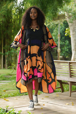 Load image into Gallery viewer, Woman in a colorful dress standing outdoors with trees and a bench in the background
