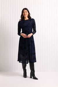 Woman wearing a dark blue  mesh and velvet dress with long sleeves, standing against a white background.