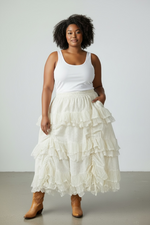 Load image into Gallery viewer, Woman wearing a white tank top and ruffled cream skirt with brown boots on a plain background
