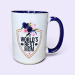 Load image into Gallery viewer, A white ceramic coffee mug with a navy blue handle, interior rim, and base. The front features a printed shield graphic with the text 'WORLD'S BEST MOM' in black, decorated with a watercolor floral design in pink, purple, and green, and a gold glitter-style border.
