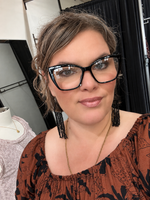 Load image into Gallery viewer, A woman with brown hair wearing black cat-eye eyeglasses, black tassel earrings, and a brown patterned top, photographed in a boutique setting.
