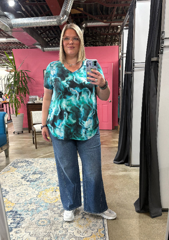 Woman taking a mirror selfie wearing a green marble-patterned shirt and blue jeans in a store.