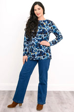 Load image into Gallery viewer, A woman modeling a long-sleeve blue abstract patterned top with blue jeans and brown ankle boots, standing against a white wall and light wood floor.
