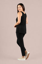 Load image into Gallery viewer, A woman modeling a black sleeveless top and black leggings with beige sneakers, shown from a side-back angle against a solid light beige studio background.
