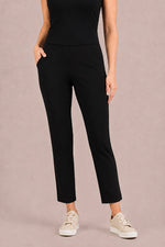 Load image into Gallery viewer, A model wearing black cropped ankle-length pants with side pockets, paired with a black top and beige sneakers, standing against a solid dusty pink studio background.
