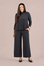 Load image into Gallery viewer, A woman modeling a dark charcoal gray two-piece set consisting of a long-sleeve mock neck top and matching wide-leg cropped pants, set against a solid light beige studio background.
