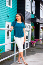 Load image into Gallery viewer, Woman in teal top and white pants standing on a sidewalk with colorful buildings in the background
